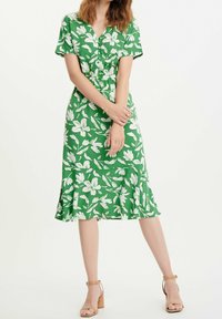 Grön blommig klänning med vita bladvärmönster, V-ringning, korta ärmar, samlad midja och utsvängd nederdel. Tillverkad av lättviktig tyg.