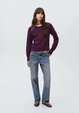 Paarse gebreide trui met lange mouwen, gecombineerd met lichtblauwe rechte jeans. Model staat met ontspannen handen en draagt bordeauxrode loafers.