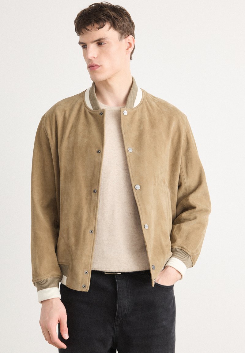 Tan suede bomber jakna z rebrastim pletenim ovratnikom in manšetami. Ima gumbe na snap in sproščen kroj, v kombinaciji s svetlim puloverjem.