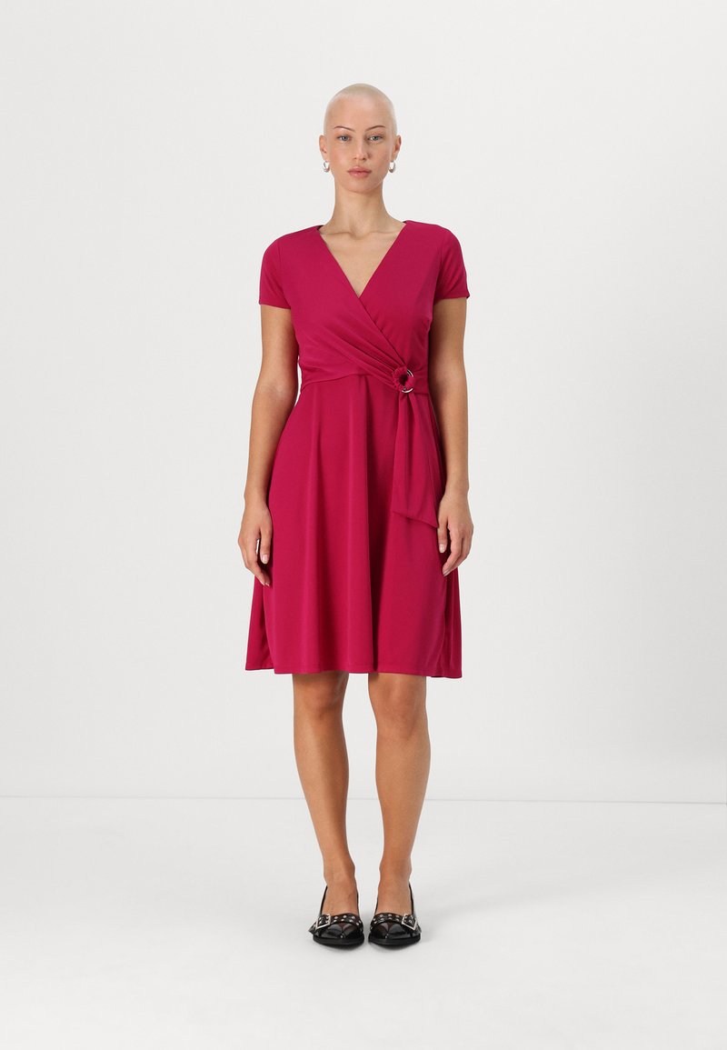 Fuksia wrap dress dengan lengan pendek, memiliki detail ikatan di pinggang dan siluet skater, dipadukan dengan sepatu hitam.