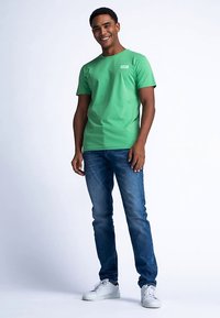 Vihreä puuvillainen t-paita, jossa on lyhyet hihat ja pyöreä pääntie, yhdistetty sinisiin farkkuihin ja valkoisiin tennareihin, seisoo vaaleaa taustaa vasten.