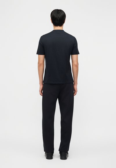 Άνδρας που στέκεται με την πλάτη, φοράει απλό μαύρο κοντομάνικο μπλουζάκι, μαύρο παντελόνι και μαύρα παπούτσια μπροστά σε απλό φωτεινό φόντο.