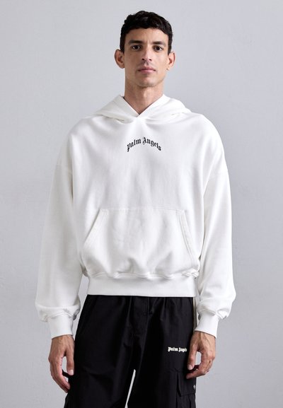 Weißer Kapuzenpullover mit Fronttasche, gesticktem "Palm Angels"-Logo, lockerer Passform und umgeschlagenen Ärmeln, kombiniert mit schwarzen Hosen.