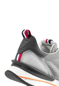 Šedá sportovní bota s texturovanou tkaninou a hladkými syntetickými akcenty. Obsahuje černou patu s logem a kontrastními červenými, bílými a modrými detaily.