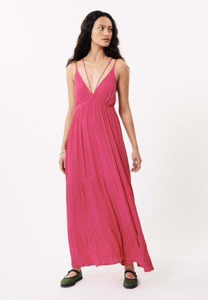 Robe maxi de couleur rose vif, dotée de fines bretelles, d'un décolleté en V profond et d'un tissu texturé à plusieurs niveaux. Associée à des chaussures vertes.