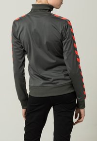 Mörkgrå sportjacka med hög krage, prydd med röda diagonala ränder längs ärmarna och en figursydd midja. Slät textur.