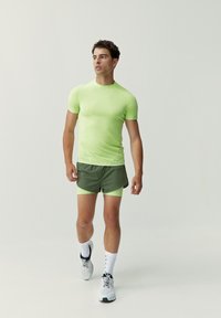 Maglietta sportiva aderente verde neon abbinata a pantaloni corti verde oliva. Il modello indossa calzini bianchi e scarpe da corsa grigio chiaro. Tessuto liscio e elastico.