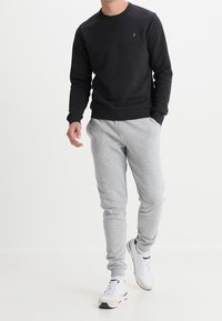 Sudadera negra con cuello redondo y un pequeño logo naranja, combinada con pantalones de chándal tapereds de color gris claro y zapatillas blancas.