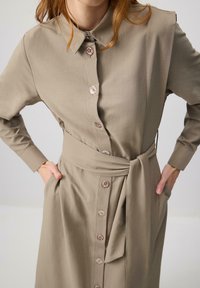 Khaki button-up dress with long sleeves, matching belt, and side pockets. Features decorative buttons and a smooth, structured fabric texture.