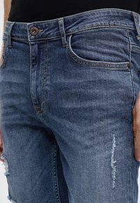 Džínové kalhoty středně modré barvy, s klasickým designem se pěti kapsami, zdobené prošitím a opotřebovaným trhlinou na stehně.