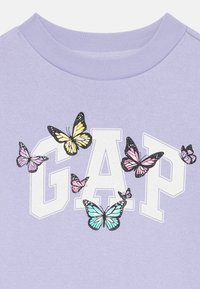 Sweatshirt à manches courtes lavande avec un logo "GAP" blanc, décoré de graphiques de papillons multicolores de différentes tailles.