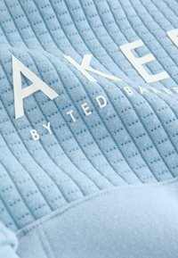 Lys blå stof med et struktureret, quiltet design. Har hvid brandingtekst "BAKER" og "by TED BAKER" på overfladen.