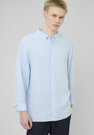 Jeune homme aux cheveux blonds courts portant une chemise bleu clair boutonnée et un jean foncé sur un fond gris uni.