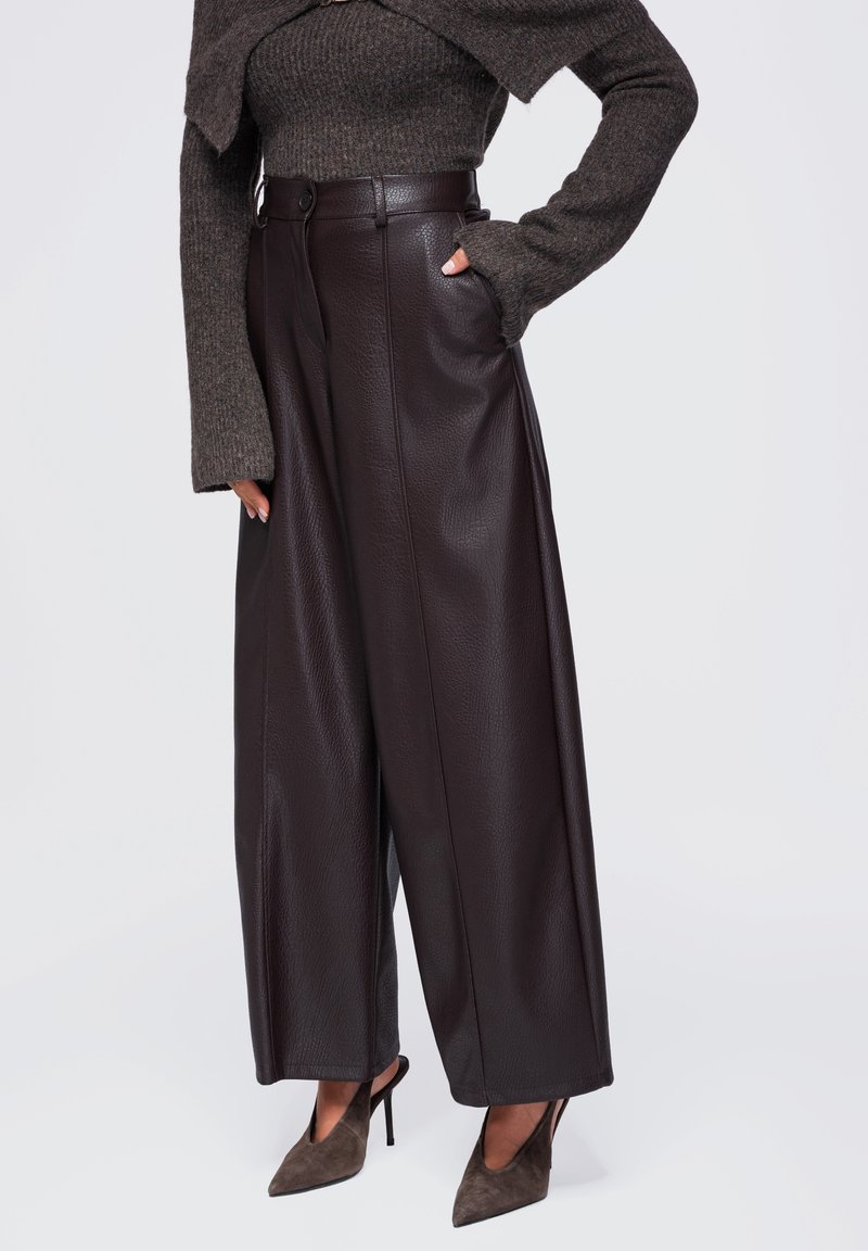 Femme portant un pantalon en cuir marron foncé à jambes larges, un pull en maille marron foncé et des chaussures à talons aiguilles pointues avec des brides à la cheville.