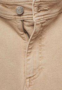 Pantaloni in denim beige in primo piano, con bottoni metallici, chiusura con zip e cuciture visibili. Tessuto morbido con una leggera lucentezza.