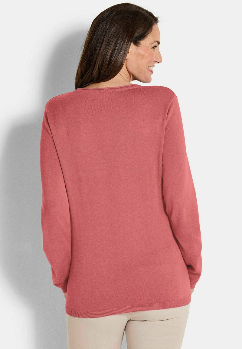 Frau mit langen braunen Haaren, die einen staubigen rosa Pullover mit langen Ärmeln und helle beige Hosen trägt, leicht gedreht, um die Rückseite des Pullovers zu zeigen.