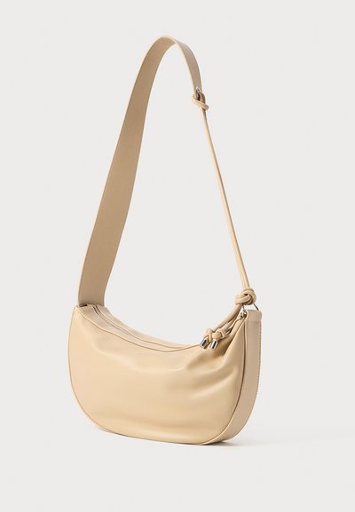 Sac à bandoulière beige avec une forme courbée, fabriqué en matériau lisse. Il dispose d'une fermeture éclair et d'un accent noué sur la sangle.