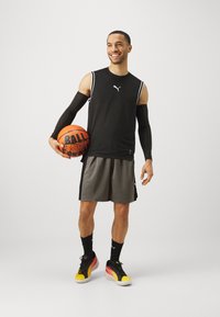 Camiseta deportiva sin mangas negra con detalles en blanco, pantalones cortos grises y zapatillas de baloncesto negras con detalles en amarillo y rojo, sosteniendo un balón de baloncesto.
