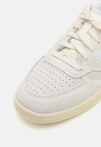 Világosszürke velúr sneaker, perforált orral, krémszínű fűzőkkel és bézs gumitalppal. Oldali logóval és varrási részletekkel rendelkezik.