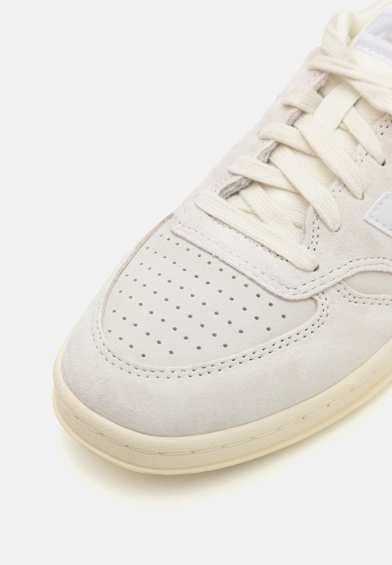 Zapatillas de ante gris claro con caja de puntera perforada, cordones crema y suela de goma beige. Presenta un логотип lateral y detalles de costura.