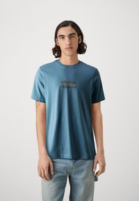 Camiseta de algodón azul con mangas cortas y cuello redondo. Presenta un gráfico rectangular en múltiples colores en la parte delantera.