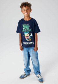 Marineblå T-skjorte med Minecraft-grafikk; ledig passform, korte ermer, med "Minecraft Movie"-tekst nederst. Kombinert med lyseblå jeans.