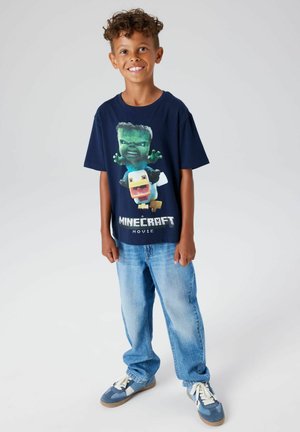 Granatowy T-shirt z grafiką Minecraft; luźny krój, krótkie rękawy, z napisem "Minecraft Movie" na dole. Połączony z jasnoniebieskimi dżinsami.