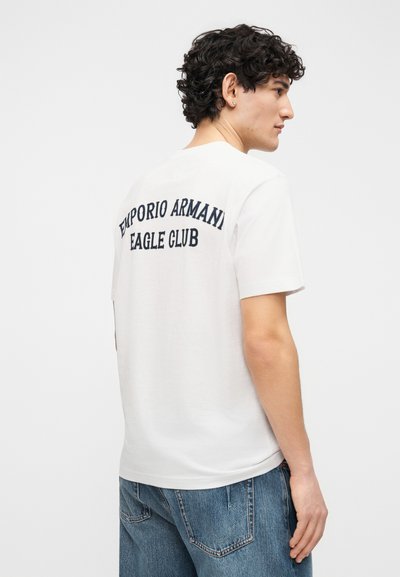 Νέος άνδρας με κατσαρά μαλλιά, φορώντας λευκό t-shirt με το κείμενο "Emporio Armani Eagle Club" στην πλάτη και μπλε τζιν παντελόνι.