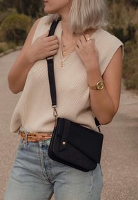 Schwarze Leder-Umhängetasche mit diagonaler Klappe, goldenen Hardware-Details, angebrachtem Schultergurt und glatter Textur. Kombiniert mit hellblauer Jeans und cremefarbener Bluse.