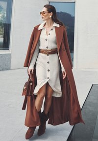 Donna con un lungo cappotto marrone su un vestito a maglia crema con bottoni, stivali marroni, che porta una borsa marrone, occhiali da sole e orecchini d'oro.