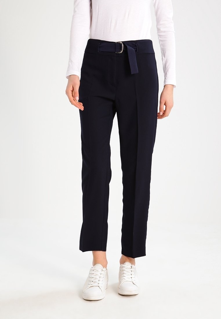 Personne vêtue d’un pantalon noir taille haute avec une ceinture en tissu, d’un haut blanc à manches longues et de baskets blanches à lacets, debout devant un fond uni.