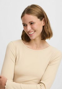 Beige långärmad topp tillverkad av ribbat tyg, med en rundad halsringning och volangärmar, som framhäver en figurnära design.