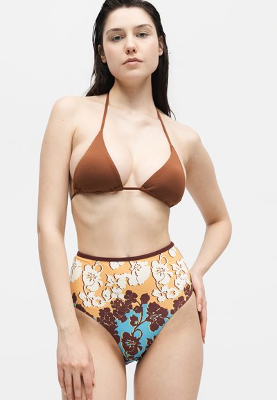 Γυναίκα που φοράει καφέ τοπ μπικίνι και ψηλόμεσα floral μπικίνι κάτω μέρη με κίτρινες, μπλε και καφέ αποχρώσεις, στέκεται σε λευκό φόντο.