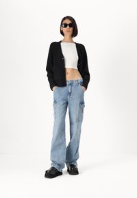 Cardigan nero a maglia con tasche frontali, crop top bianco, jeans cargo azzurri chiari con tasche laterali e scarpe platform nere.