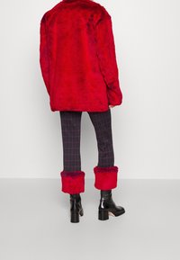 Veste en fausse fourrure rouge au toucher pelucheux, portée par-dessus un pantalon à carreaux sombres. Les bottes présentent un talon épais et des accents de fourrure autour des chevilles.