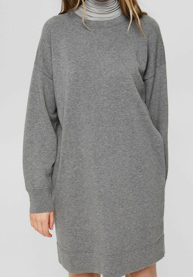 Femme portant une longue robe-pull grise avec épaules tombantes et poignets côtelés par-dessus un col roulé gris clair.