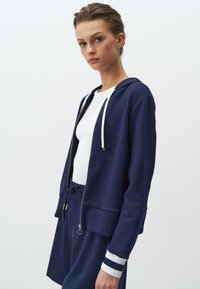 Felpa con zip color blu navy con lacci bianchi, indossata sopra una maglietta bianca. Presenta polsini a coste con strisce blu e bianche. Tessuto morbido e casual.