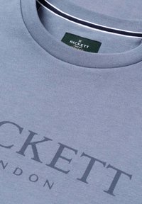 Maglietta azzurro chiaro con scollo girocollo e la scritta "Hackett London" stampata in grandi lettere scure sul petto, con etichetta nera sul colletto.