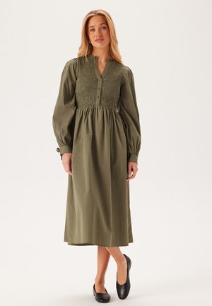 Olijfgroene jurk met lange mouwen, een smock-borstvoesite en knopenlijst, een vloeiende rok en taps toelopende mouwen, gecombineerd met zwart platte schoenen.