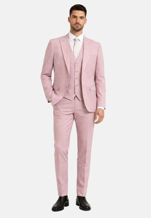 Homme portant un costume trois pièces rose clair avec des chaussures habillées noires et une chemise blanche avec une cravate de couleur claire, debout avec une main dans la poche.