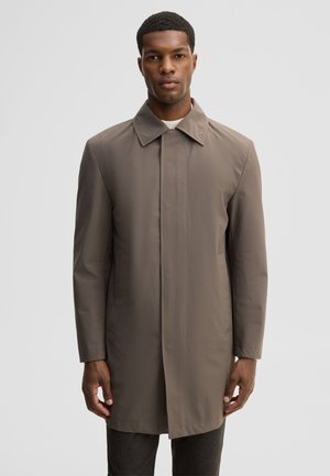 Man wearing a taupe knee-length coat with a collar and concealed front buttons, standing against a plain light gray background.