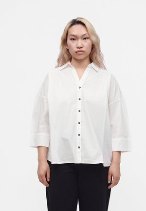 Ung person med langt blondt hår står med front mod kamera iført en løs hvid skjorte med knapper og sorte bukser mod en ensfarvet hvid baggrund.