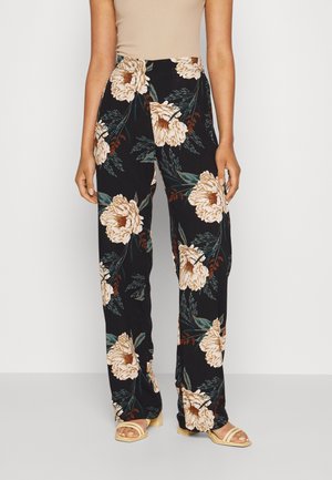 Frau trägt schwarze, hoch taillierte, weit geschnittene Hose mit großem beigem Blumenmuster und beigefarbene Sandalen mit offenen Zehen, steht vor einem einfarbigen Hintergrund.