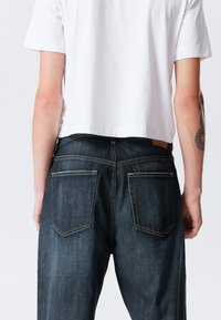 Jeans de ganga azul escuro com costura branca e dois bolsos traseiros, usados com uma t-shirt branca curta. Textura lisa, design casual.