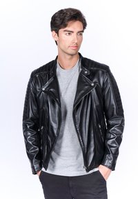 Giacca da motociclista in pelle nera con dettagli trapuntati sulle spalle, cerniere argentate e vestibilità slim, indossata sopra una maglietta grigia e pantaloni neri.