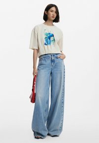 T-shirt beige surdimensionné avec un motif graphique bleu, associé à un jean en denim léger bleu large. Le modèle tient un sac rouge.