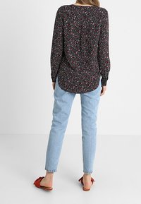 Blouse noire à manches longues avec un motif floral, associée à un jean bleu clair et des sandales plates rouges. La blouse présente un ourlet arrondi.