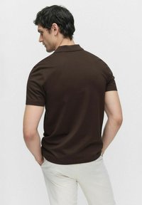 Bruine polo met korte mouwen en een omgeslagen kraag, gemaakt van gladde stof. Het model staat met de rug naar de camera, waardoor een aansluitend ontwerp en neutrale broek zichtbaar zijn.