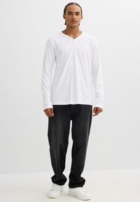 Camisa henley de manga larga blanca con abertura de tres botones, combinada con jeans negros y zapatillas blancas. El modelo se encuentra de pie frente a un fondo neutro.