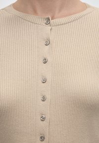 Κοντινό πλάνο μπεζ ζακάρ ζακέτας με μικρά στρογγυλά κουμπιά κλειδωμένα κατά μήκος του κέντρου του μπροστινού μέρους στο λαιμό.